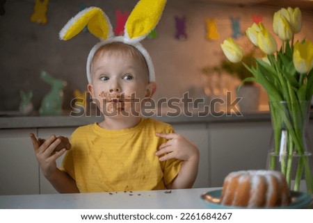 A cute little boy wearing bunny ears on Easter day is eating a chocolate Easter bunny. A child plays egg hunt for Easter. Charming child celebrates Easter at home. Royalty-Free Stock Photo #2261684267