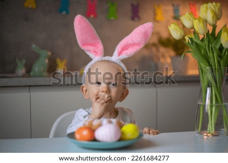 happy cute little baby girl wearing bunny ears on easter day sitting at the table in the kitchen and eats a birthday cake.