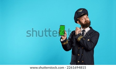 Positive aircrew captain showing phone with greenscreen, holding smartphone with isolated display and chroma key template. Young airplane pilot in uniform looking at mockup copyspace screen.