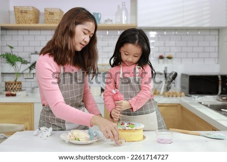 Happy lovely asian single mom and cute daughter in apron having fun with teaching and decorating homemade cake in the kitchen. Family lifestyle cooking with education concept.