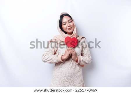 A happy young Asian Muslim woman wearing a hijab feels romantic shapes heart gesture expressing tender feelings and holding a red heart-shaped paper