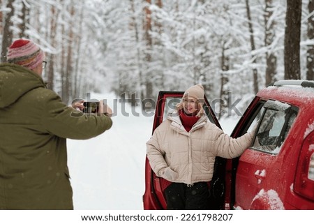 Smiling mature woman in winterwear standing by open door of car and posing for her husband with smartphone taking picture of her