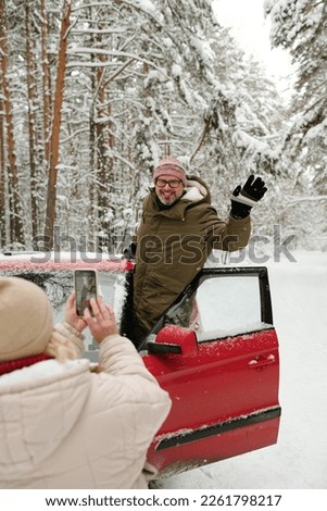 Cheerful mature male hiker in winterwear waving hand while standing in door of red car and posing for his wife with smartphone