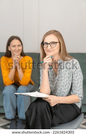 Smiling female psychologist with glasses, leaning her hand on her chin, looks into the camera, consulting a young woman in the office interior. Therapy, psychological support, professional help