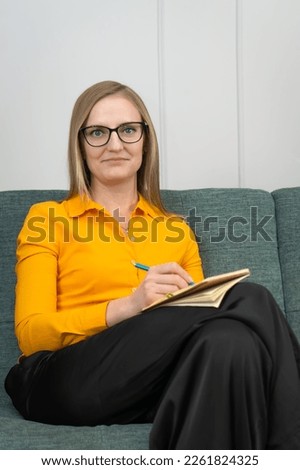 Beautiful young psychotherapist takes notes, looks into camera and smiles while sitting on couch in office. Portrait beautiful female psychologist with glasses, posing with clipboard in room