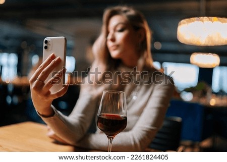 Selective focus of blurred young woman sitting at table with glass of red wine using mobile phone live on social media, writing stories, taking selfies and blogs at restaurant with dark interior.
