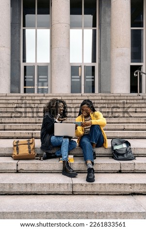 Stock photo of black cheerful friends using laptop and cellphone while sitting in stairs.