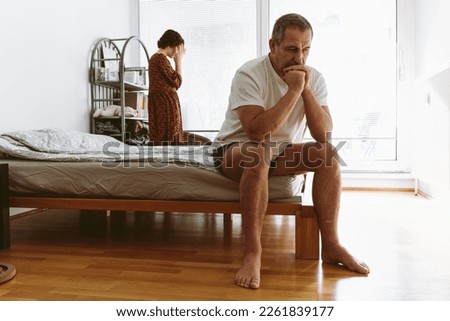 middle-aged man sits at end of bed and holds head after an argument with wife standing at window, side view. Royalty-Free Stock Photo #2261839177