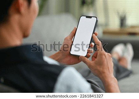 Young man resting on couch and checking social media on moblie phone. Over shoulder closeup view Royalty-Free Stock Photo #2261841327