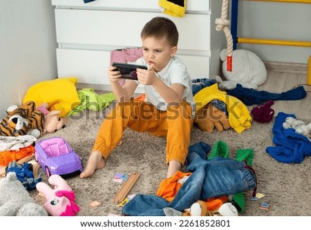 boy (5-6 years old) in bedroom, uses phone, plays phone games in dirty children's room, child plays among lot of toys at home,lot of toys and things scattered on floor of the room, a dirty house.
