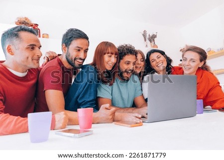 group of seven happy friends watching online streaming together on laptop in the kitchen of a house