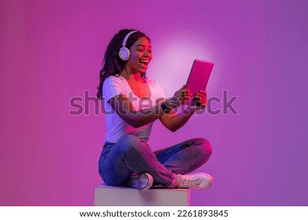 Virtual Content. Excited Black Female Relaxing With Digital Tablet While Sitting On Big Cube In Neon Light, Young African American Woman Looking At Glowing Device Screen And Smiling, Copy Space