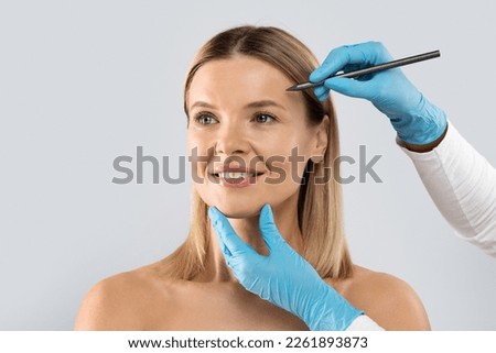 Middle aged attractive blonde woman attending aesthetic clinic, unrecognizable doctor plastic surgeon making pre surgery marks on adult lady face before beauty procedure, copy space Royalty-Free Stock Photo #2261893873