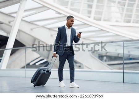 Portrait Of Handsome Black Businessman Walking With Suitcase In Airport And Using Smartphone, Young African American Man In Suit Browsing Internet On Cellphone While Going To Flight Gate, Copy Space Royalty-Free Stock Photo #2261893969
