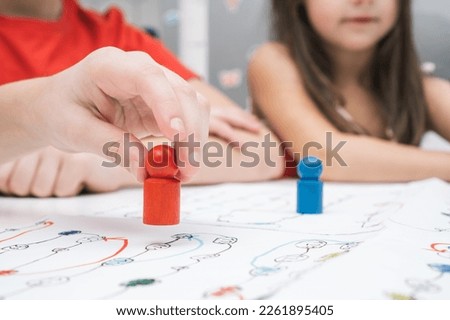 Little friends play board game on paper map with dice and chips at table. Hand of player hold red figure closeup, selective focus. Home leisure, playtime. Entertaining interesting table game. Royalty-Free Stock Photo #2261895405