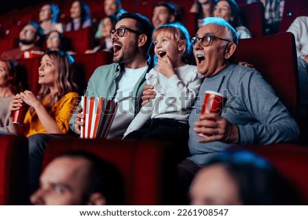 A grandfather and his son accompanied with his granddaughter are in the movie theater watching and exciting film together while enjoying beverages and popcorn. Royalty-Free Stock Photo #2261908547