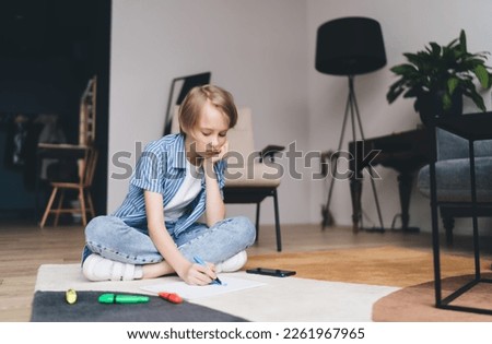 Bored boy in casual clothes leaning on hand and drawing picture with felt pen on paper while sitting cross legged on floor at home