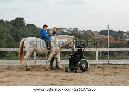 Boy with disabilities riding a horse while having an equine therapy session with an instructor. Royalty-Free Stock Photo #2261973255