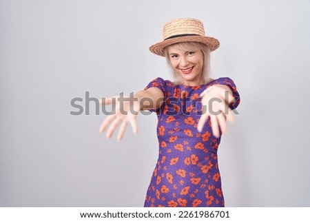 Young caucasian woman wearing flowers dress and summer hat smiling cheerful offering hands giving assistance and acceptance. 