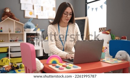Young beautiful hispanic woman preschool teacher using laptop sitting on table at kindergarten Royalty-Free Stock Photo #2261995787