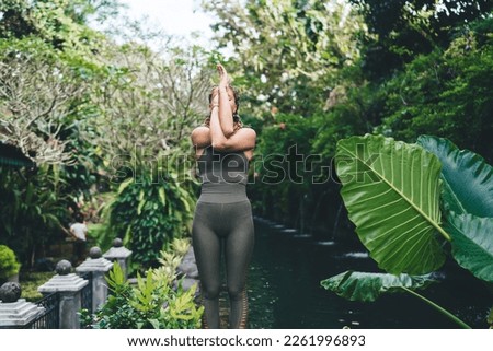 Fit female athlete in sportswear meditating and practicing Pranayama breathing exercise with entangled hands while relaxing in tropical garden on vacation Royalty-Free Stock Photo #2261996893