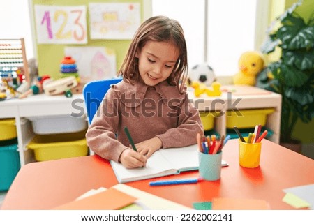 Adorable hispanic girl preschool student sitting on table writing on notebook at kindergarten Royalty-Free Stock Photo #2262002141