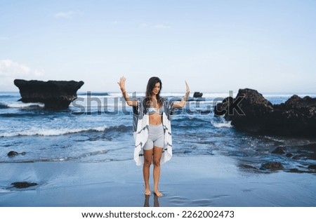 Full length of young fit ethnic barefooted female tourist with long hair standing on wet sandy seashore near rocky formations with closed eyes and raised arms while meditating during yoga