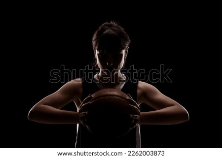 Female basketball player. Beautiful girl holding ball. Side lit half silhouette studio portrait against black background.