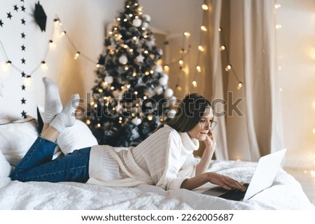 young female software tester in casual clothes lying on bed and working on laptop while looking at screen and working on testing jobs for remote company in decorated room with Christmas tree