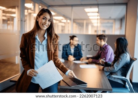 Portrait of young happy businesswoman during a meeting in the office looking at camera. Her colleagues are in the background. 