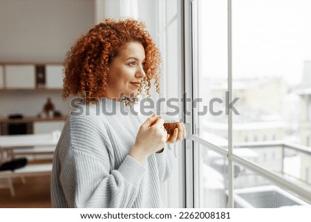 Side view indoor portrait of young attractive woman with red curly hair looking through window holding transparent cup of hot tea, admiring winter cityscape, dressed in stylish sweater Royalty-Free Stock Photo #2262008181