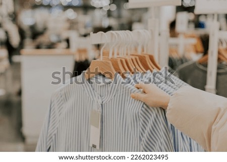 The hand of a young unrecognizable caucasian girl chooses women's blue-and-white striped shirts hanging on a hanger on a stand in a clothing boutique, while shopping, close-up side view.