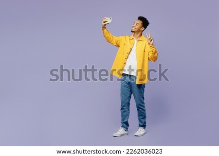 Full body young man of African American ethnicity wear yellow shirt t-shirt doing selfie shot on mobile cell phone post photo on social network isolated on plain pastel light purple background studio
