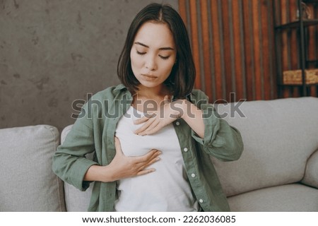 Young serious calm woman of Asian ethnicity wears casual clothes put hand on chest check breast sits on grey sofa couch stay at home hotel flat rest relax spend free spare time in living room indoor Royalty-Free Stock Photo #2262036085