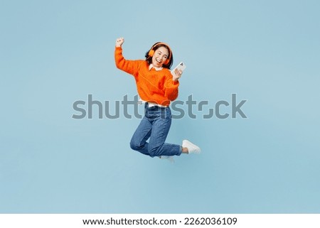Full body young woman of Asian ethnicity wear orange sweater glasses headphones listen to music raise up hands dance jump high use mobile cell phone isolated on plain pastel light blue cyan background Royalty-Free Stock Photo #2262036109