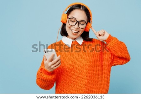 Young woman of Asian ethnicity wear orange sweater glasses headphones listen to music raise up hands dance have fun on party use mobile cell phone isolated on plain pastel light blue cyan background Royalty-Free Stock Photo #2262036113