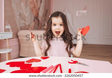 a little girl makes Valentine's Day cards using colored paper, scissors and pencil, sitting at a table in a cozy room. Royalty-Free Stock Photo #2262050457