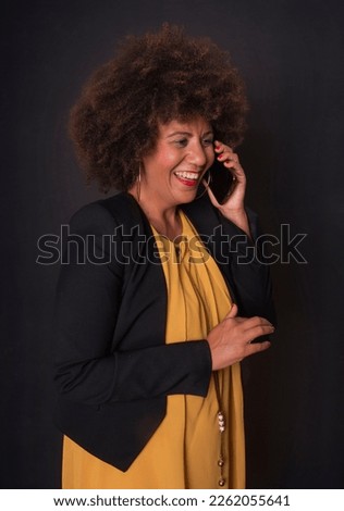 woman of color, brunette with afro hair, talking on the phone laughing, with a positive and cheerful attitude, wearing a yellow dress with a dark jacket.
