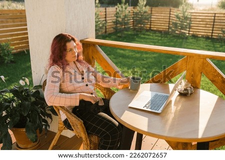 Woman working with laptop outdoor in summer terrace with black kitten. Girl and cat watching video on computer in the evening. Working from home, business, pet and lifestyle concept.