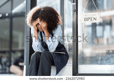 African barista woman in a barista uniform holding file showing stress and frustration sitting the entrance of a coffee shop there are no customers at the store Royalty-Free Stock Photo #2262072983
