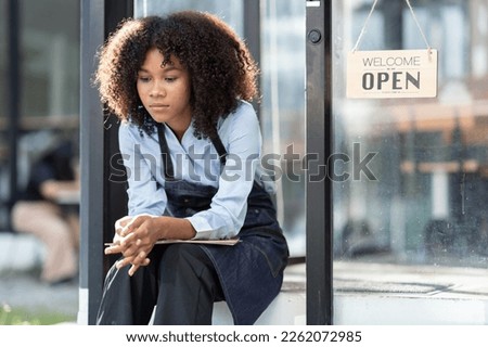 African barista woman in a barista uniform holding file showing stress and frustration sitting the entrance of a coffee shop there are no customers at the store Royalty-Free Stock Photo #2262072985