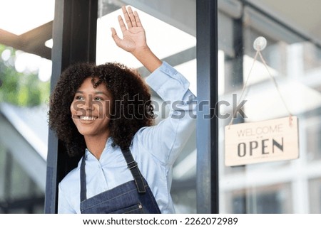 African young woman starting a small business working in a cafe A coffee shop worker stands in front of the store opening an open sign and greets customers with happy smiles.
