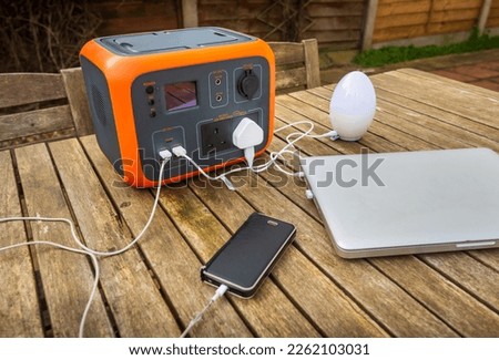 Portable power station solar electricity generator outdoors on wooden table with laptop, mobile phone and lamp electronic devices charging. Wireless charging lithium battery backup for use off grid. Royalty-Free Stock Photo #2262103031