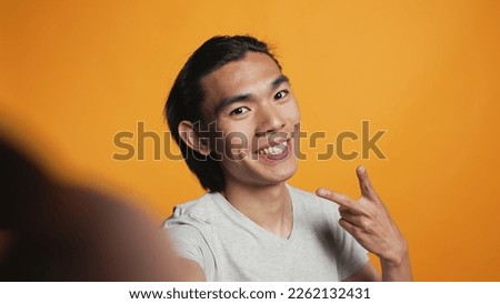 POV of asian smiling man taking pictures on camera, acting funny and silly over orange background. Cheerful casual model posing for photos in studio, smiling and being happy. Carefree hipster.