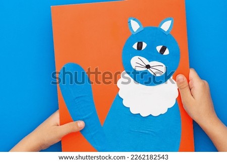 Top view of child hands holding paper craft cat on red sheet on blue background