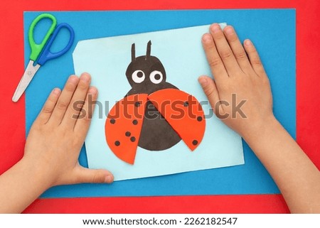 Top view of child hands with papercraft ladybug made by child and scissors on multicolor sheets background