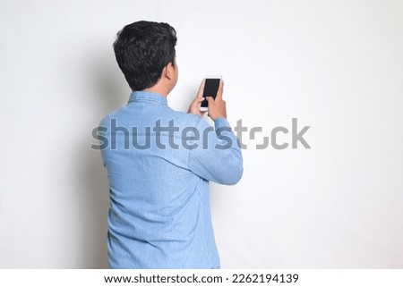 Back view portrait of excited Asian man in blue shirt holding a mobile phone and taking picture. Isolated image on white background