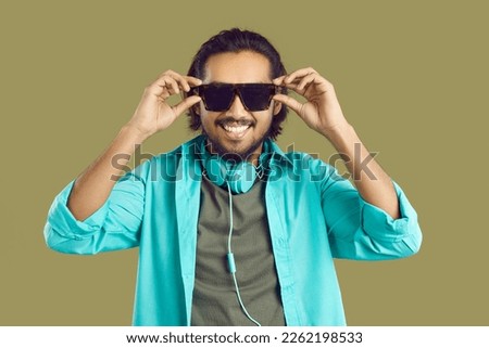Portrait of cool smiling Indian man in modern stereo headphones around his neck and in sunglasses. Close up of cheerful stylish ethnic guy wearing headphones and wearing glasses on khaki background.