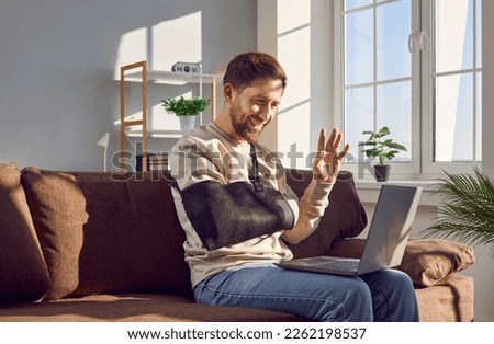 Smiling young man with broken arm making video call using laptop. Cheerful handsome man wearing arm splint sitting on sofa at home and waving his hand in greeting having meeting online Royalty-Free Stock Photo #2262198537