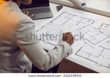 Female architect sitting at her office desk and working with blueprints, cadastral maps and city plans. Young African American woman studying information and developing a new architectural project Royalty-Free Stock Photo #2262198541